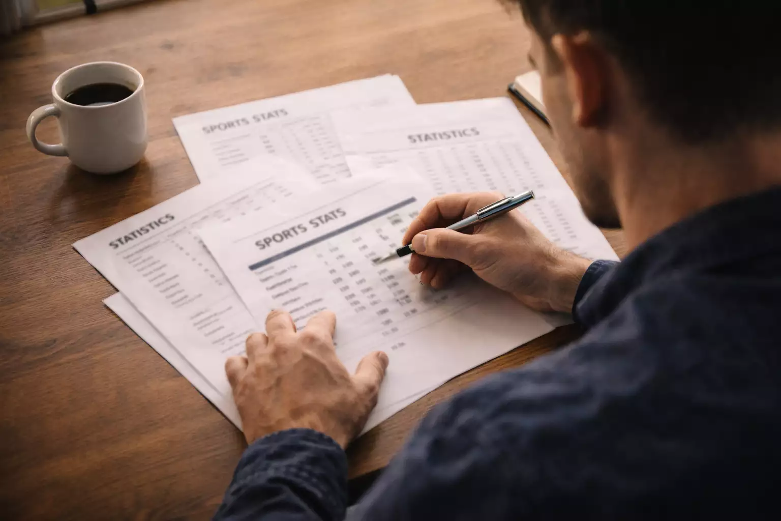 Persona che confronta diversi fogli con numeri su una scrivania ordinata, luce naturale dalla finestra